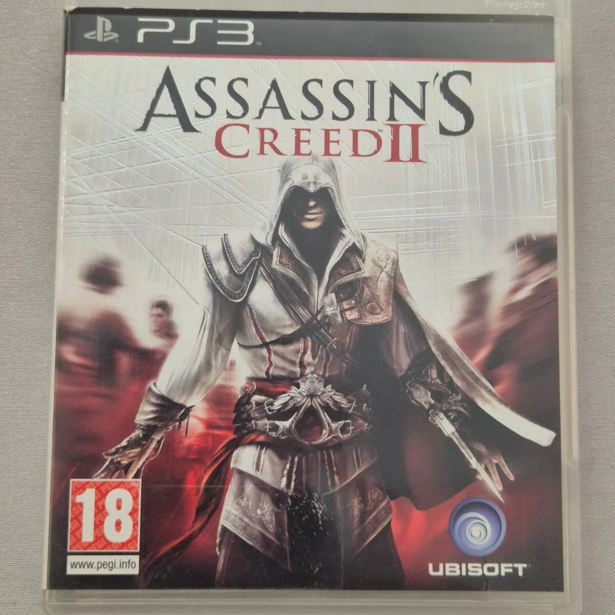 Assassin's Creed II video game for PlayStation 3 in a clear plastic case, showing the character Ezio with the Ubisoft and PEGI 18 logos on the cover, photographed on a neutral gray background.