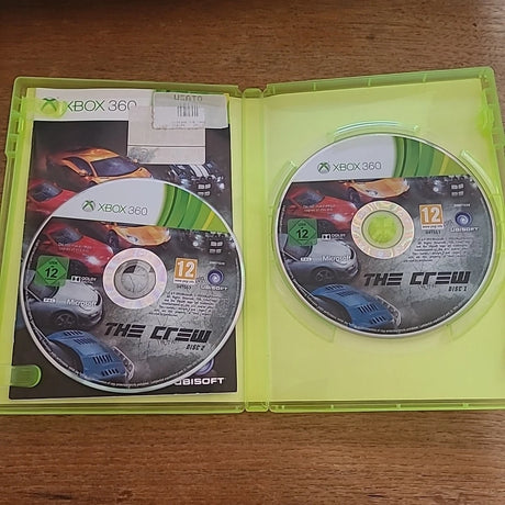 Open Xbox 360 game case for The Crew, showing the game disc on the right and the insert card on the left, placed on a wooden surface.