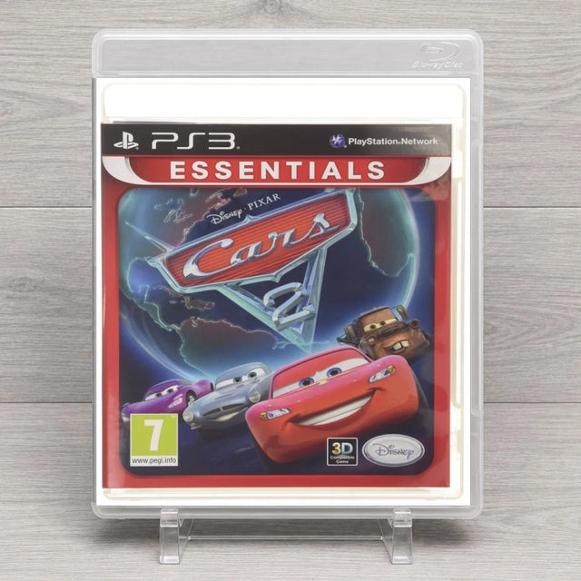 PS3 Disney Pixar Cars 2 Essentials game case in a clear plastic protective stand, displayed on a light gray wood-textured surface.