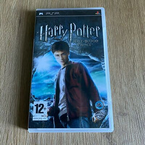A PSP game case for Harry Potter and the Half-Blood Prince, featuring artwork of Harry wearing his red robe against a blue magical background, photographed on a light wood surface.